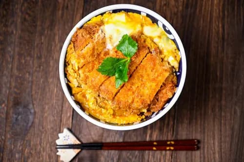 Bol de katsudon japonais avec tonkatsu de porc pané croustillant, œuf mollet, oignons fondants et riz blanc, servi dans un bol à donburi traditionnel.