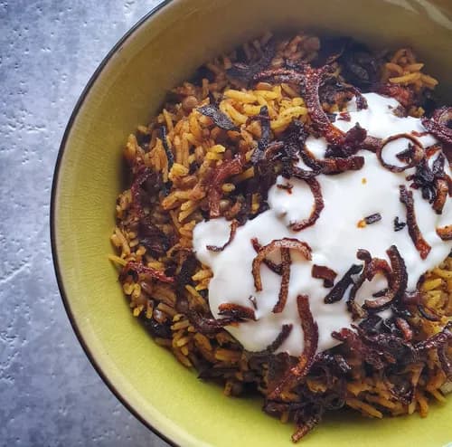 Assiette de moujadara libanaise garnie d'oignons caramélisés brun acajou sur un lit de riz et de lentilles, servie avec du yaourt nature et une salade de concombre.