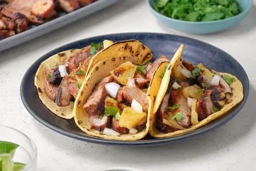 Tacos al pastor garnis de porc mariné à l'achiote grillé, ananas caramélisé, oignon et coriandre, sur tortillas de maïs chaudes