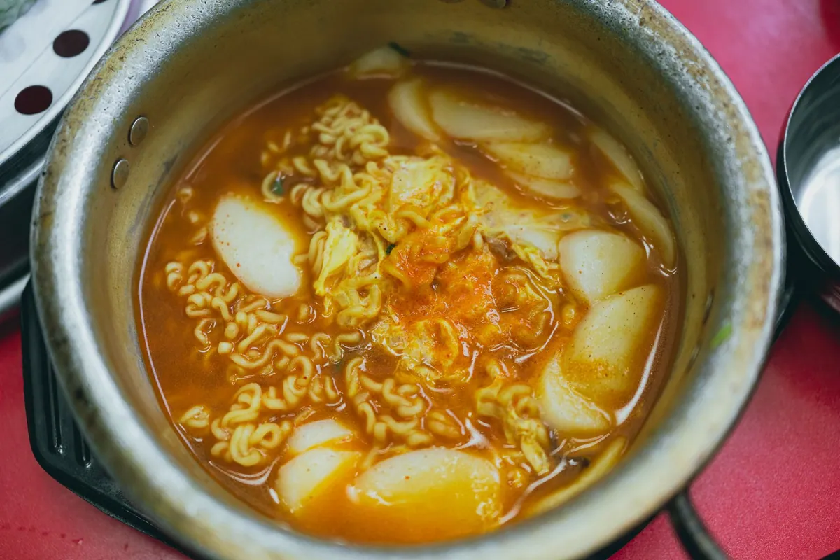 Tteokbokki coréen, gâteaux de riz nappés d'une sauce rouge au gochujang, avec galettes de poisson, oignons nouveaux et œuf dur, servi fumant dans une poêle.