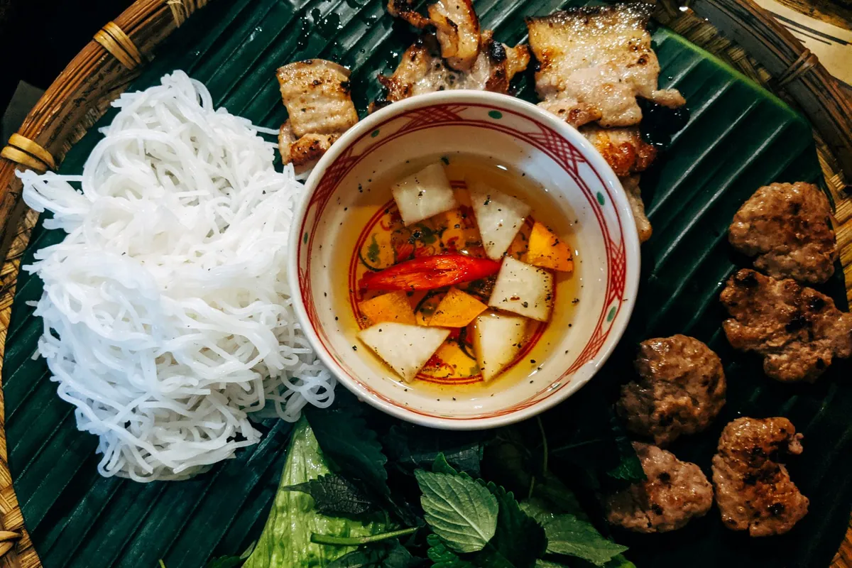 Bún chả hanoïen : galettes et tranches de porc grillées caramélisées trempées dans un bol de nước chấm tiède, servies avec vermicelles de riz et un plateau d'herbes fraîches.