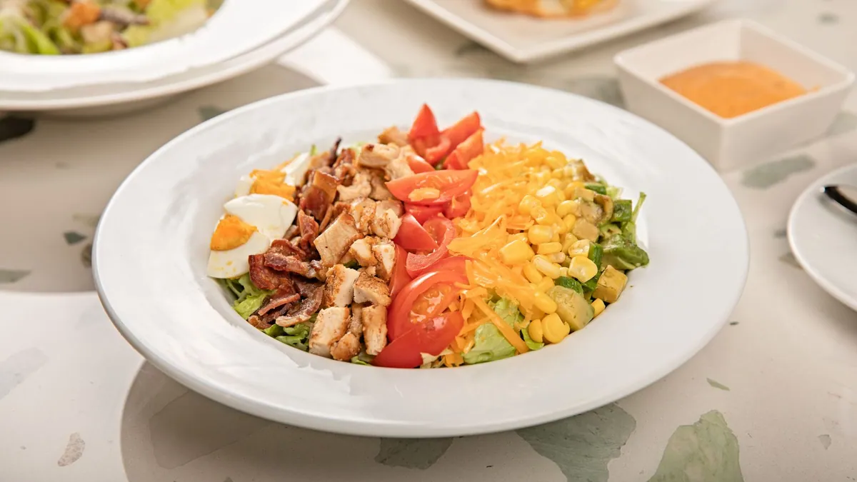 Cobb salad dressée en rangées colorées de poulet, œuf dur, bacon, avocat, tomates cerises et fromage bleu sur un lit de laitue romaine, avec une vinaigrette moutardée.