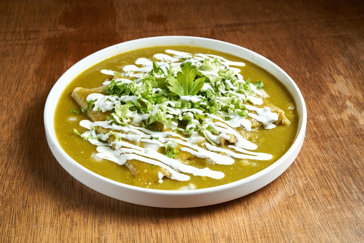 Enchiladas verdes gratinées nappées de salsa verte de tomatilles, garnies de crema et de queso fresco émietté, parsemées de coriandre et d'oignon rouge, servies dans un plat en céramique.