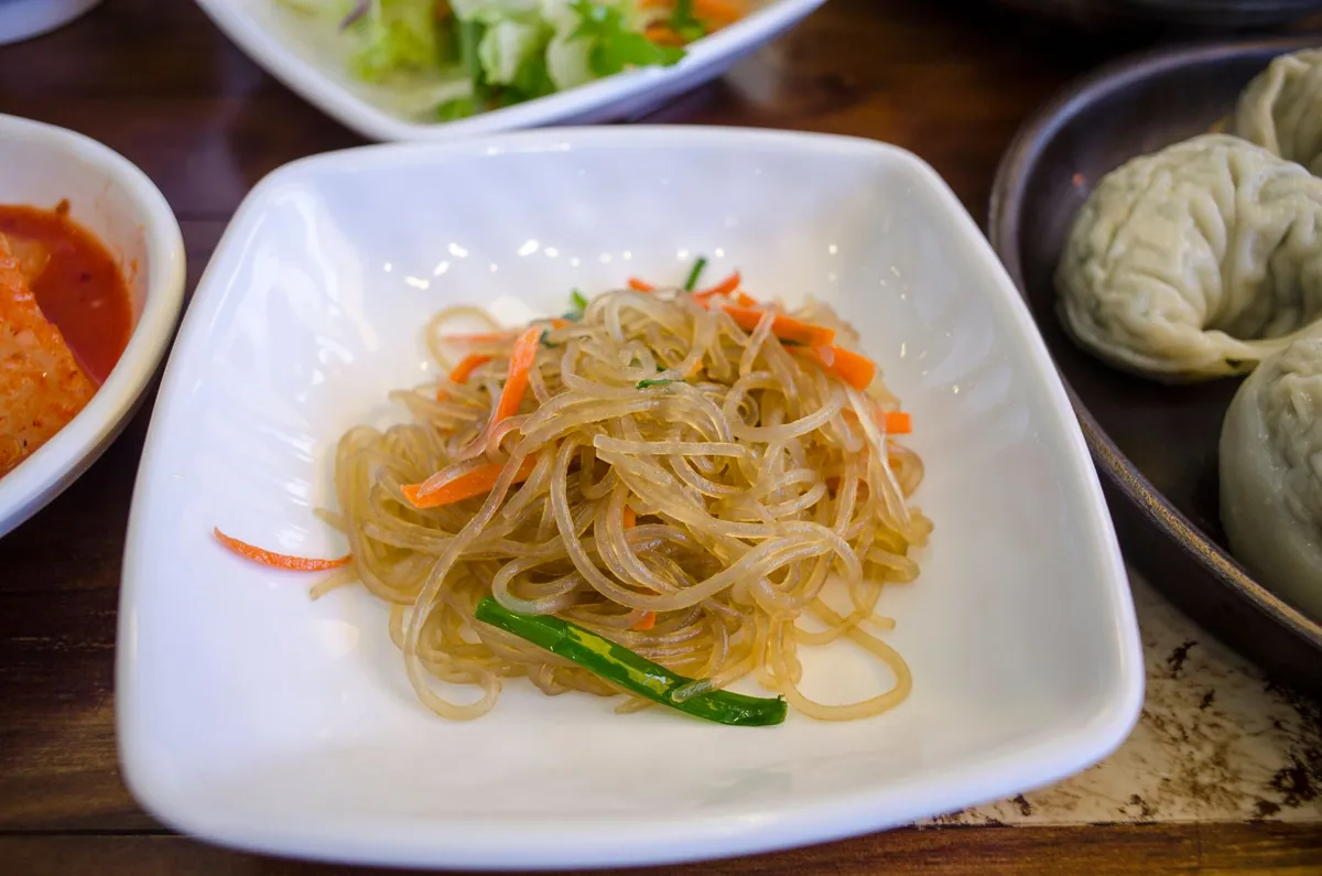 Japchae coréen : nouilles de patate douce sautées avec carotte, poivron, oignon et épinards, parsemées de graines de sésame, dans un grand plat de service.