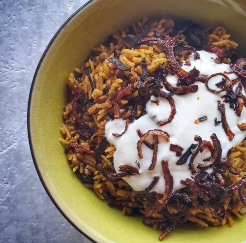 Assiette de moujadara libanaise garnie d'oignons caramélisés brun acajou sur un lit de riz et de lentilles, servie avec du yaourt nature et une salade de concombre.
