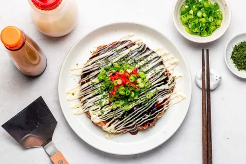 Okonomiyaki doré à la plancha, nappé de sauce et de mayonnaise en zigzag, parsemé d'aonori et de copeaux de bonite séchée qui ondulent sous la chaleur