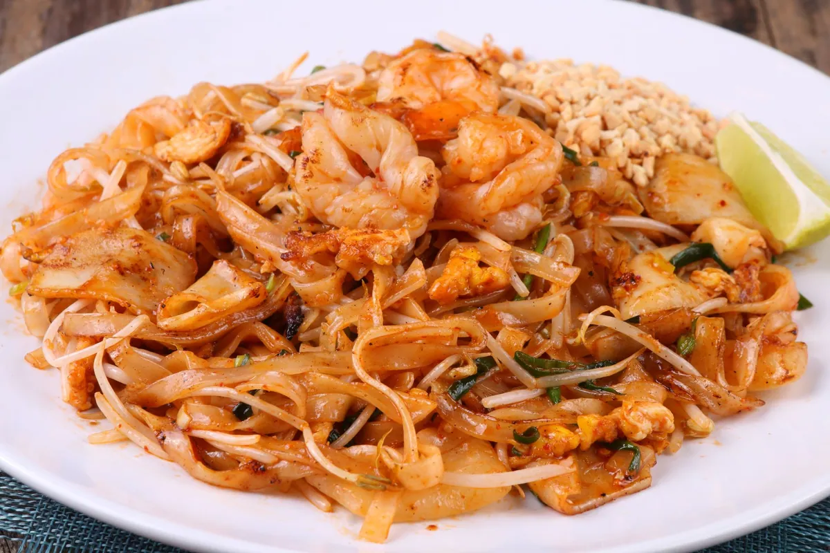 Pad thaï aux crevettes dans une assiette creuse, nouilles de riz sautées avec crevettes, tofu doré et œuf, parsemées de cacahuètes concassées, servi avec un quartier de citron vert.