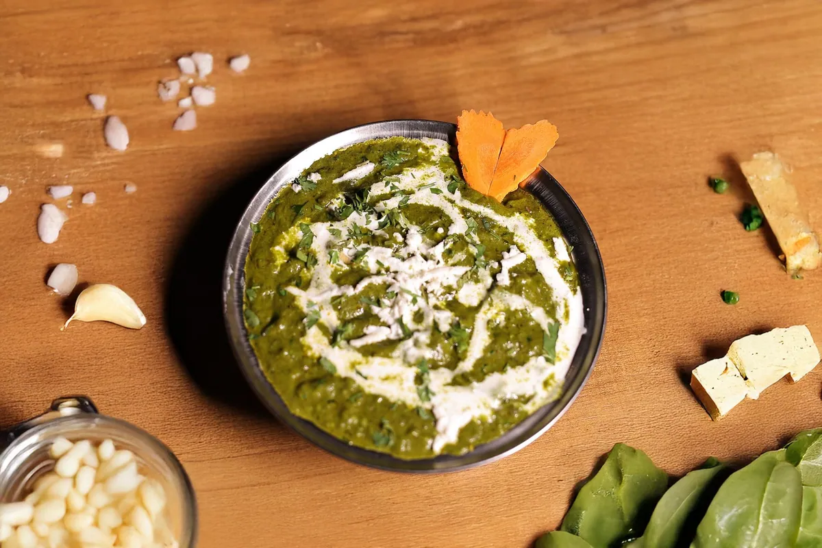 Palak paneer dans un bol en inox : purée d'épinards vert profond nappée d'un filet de crème et parsemée de coriandre ciselée, entourée de cubes de paneer, de gousses d'ail et de pousses d'épinards frais.