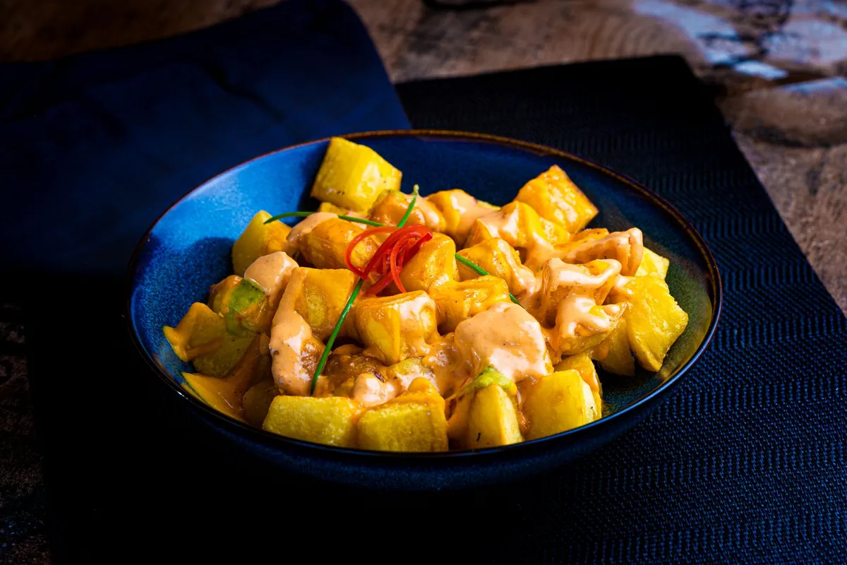 Patatas bravas dans une cazuela en terre cuite, cubes de pommes de terre rôtis et croustillants nappés de sauce brava tomatée et fumée, parsemés de paprika.
