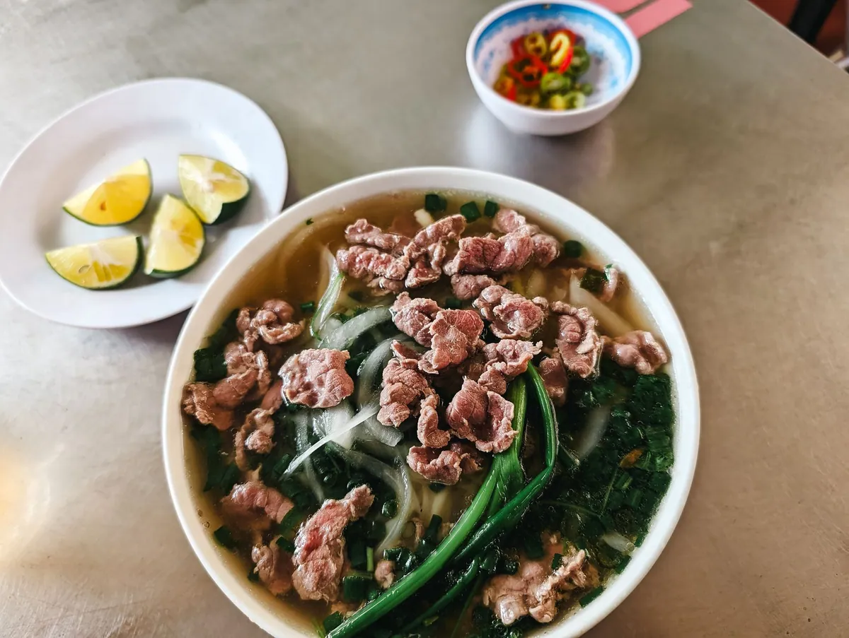 Bol de phở bò fumant avec bouillon clair, nouilles de riz plates, fines lamelles de bœuf, germes de soja, herbes fraîches et quartiers de citron vert.