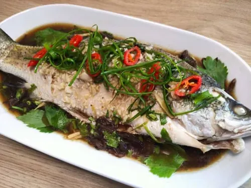 Poisson entier thaïlandais cuit à la vapeur, nappé d'une sauce au citron vert, à l'ail et au piment oiseau, parsemé de coriandre fraîche et d'ail doré croustillant, servi avec du riz jasmin.