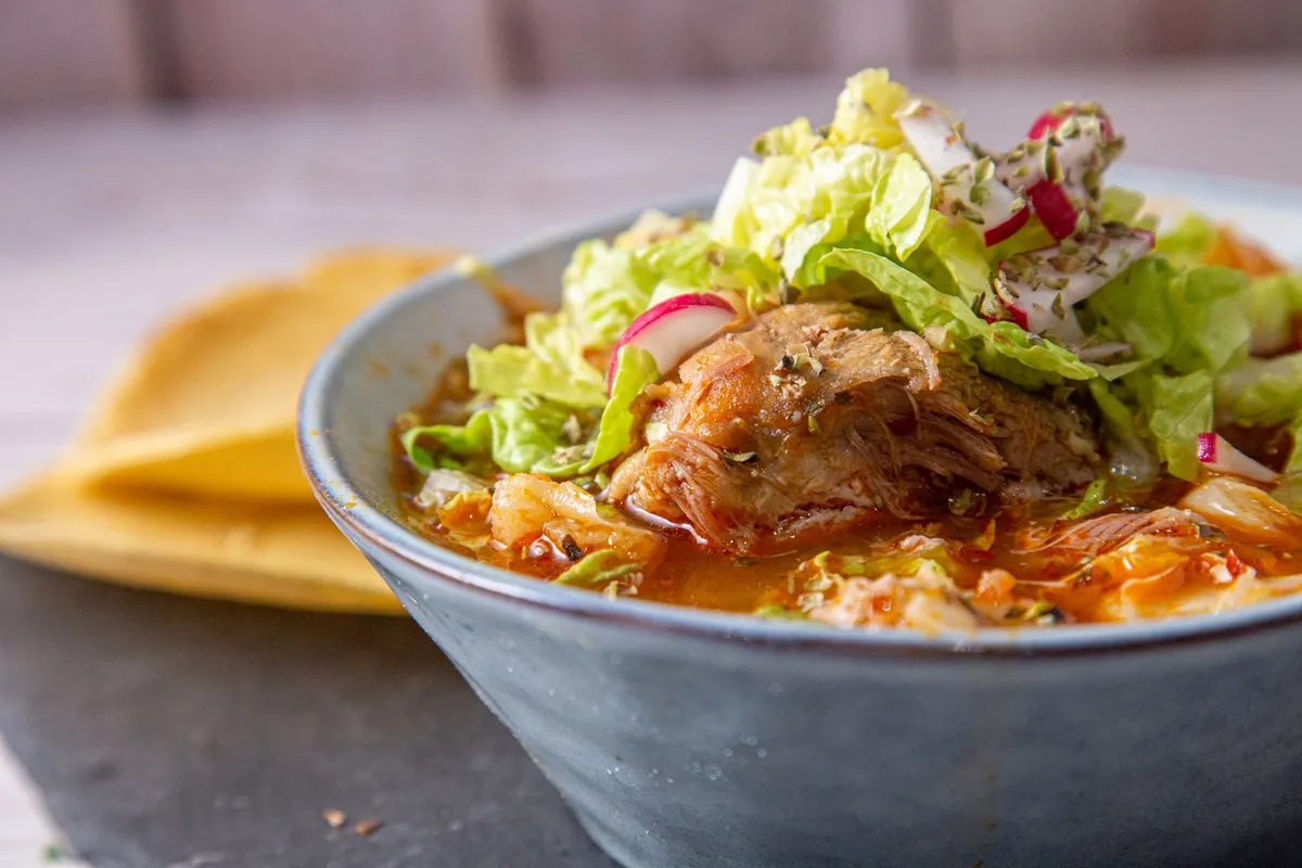 Bol de pozole rojo mexicain avec porc effiloché, maïs pozole et bouillon rouge aux piments, garni de chou émincé, radis, oignon et citron vert, servi avec des tostadas