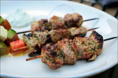 Brochettes de souvlaki de porc grillées et dorées, servies avec tzatziki maison, pita tiède, oignon rouge et quartiers de citron sur une planche.