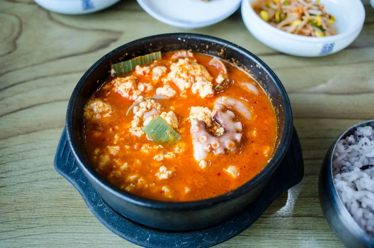 Ttukbaegi de sundubu jjigae bouillonnant, ragoût rouge de tofu soyeux, palourdes et crevettes, œuf cru à la surface, servi avec un bol de riz blanc.